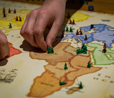 Op de afbeelding plaatsen de spelers met hun handen kleine soldaatjes op een wereldkaart die lijkt op een strategisch bordspel. Er zijn verschillende continenten te zien met gekleurde gebieden en fiches die controle of legers aangeven.