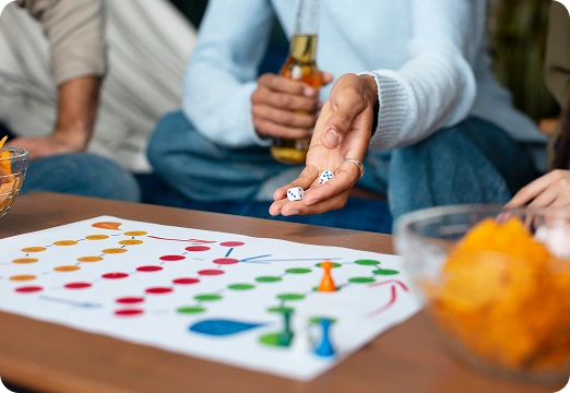 Een man speelt een bordspel met vrienden, met de dobbelstenen in de ene hand en een biertje in de andere, en rondom liggen hapjes.