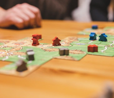 Op de afbeelding is een bordspel te zien met uitgelegde landkaarttegels en kleurrijke figuurtjes, dat lijkt op Carcassonne. Ernaast is de hand van een speler te zien die waarschijnlijk zijn zet doet.