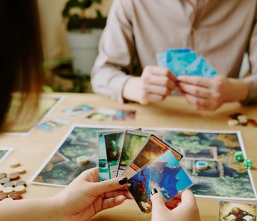 Twee mensen spelen een kaartspel aan tafel en bekijken hun kaarten aandachtig. De sfeer is rustig en geconcentreerd, met een huiselijke gezelligheid en strategische elementen tijdens het spel.