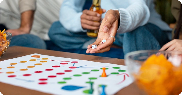 Een man speelt een bordspel met vrienden, met de dobbelstenen in de ene hand en een biertje in de andere, en rondom liggen hapjes.